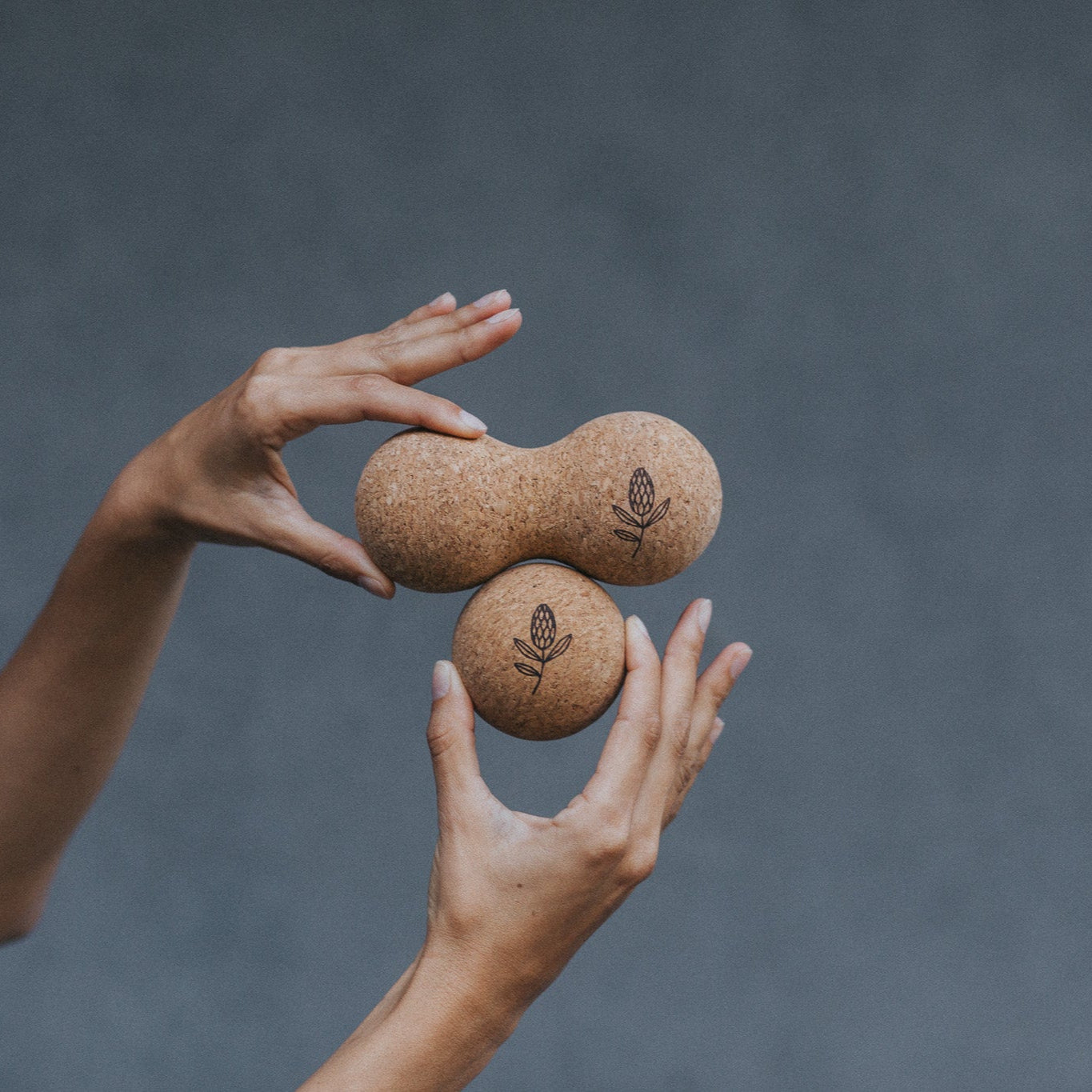 Two hands holding two cork massage rollers against a grey background