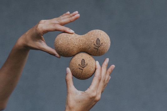Two hands holding two cork massage rollers against a grey background