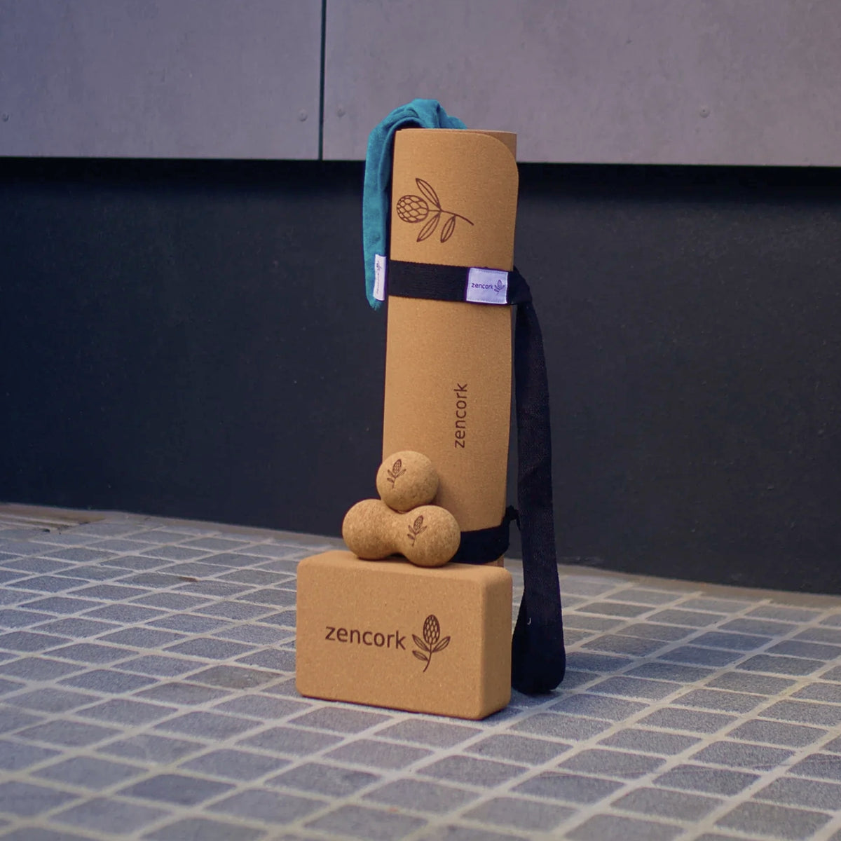 A cork yoga set including a rolled tan cork mat with a teal fabric cover, three small cork massage balls, and a tan cork block, all branded 'zencork' with a botanical logo, arranged on a tiled floor against a dark wall.