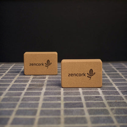 Two rectangular cork blocks with the brand name 'zencork' and a small leaf logo printed on them, displayed on a gray grid surface against a dark background.