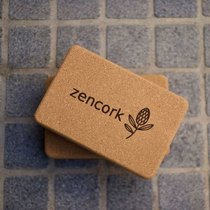 Two cork yoga blocks stacked on top of each other, featuring the 'zencork' brand name and logo printed on the top block. The blocks are placed on a gray tiled surface.