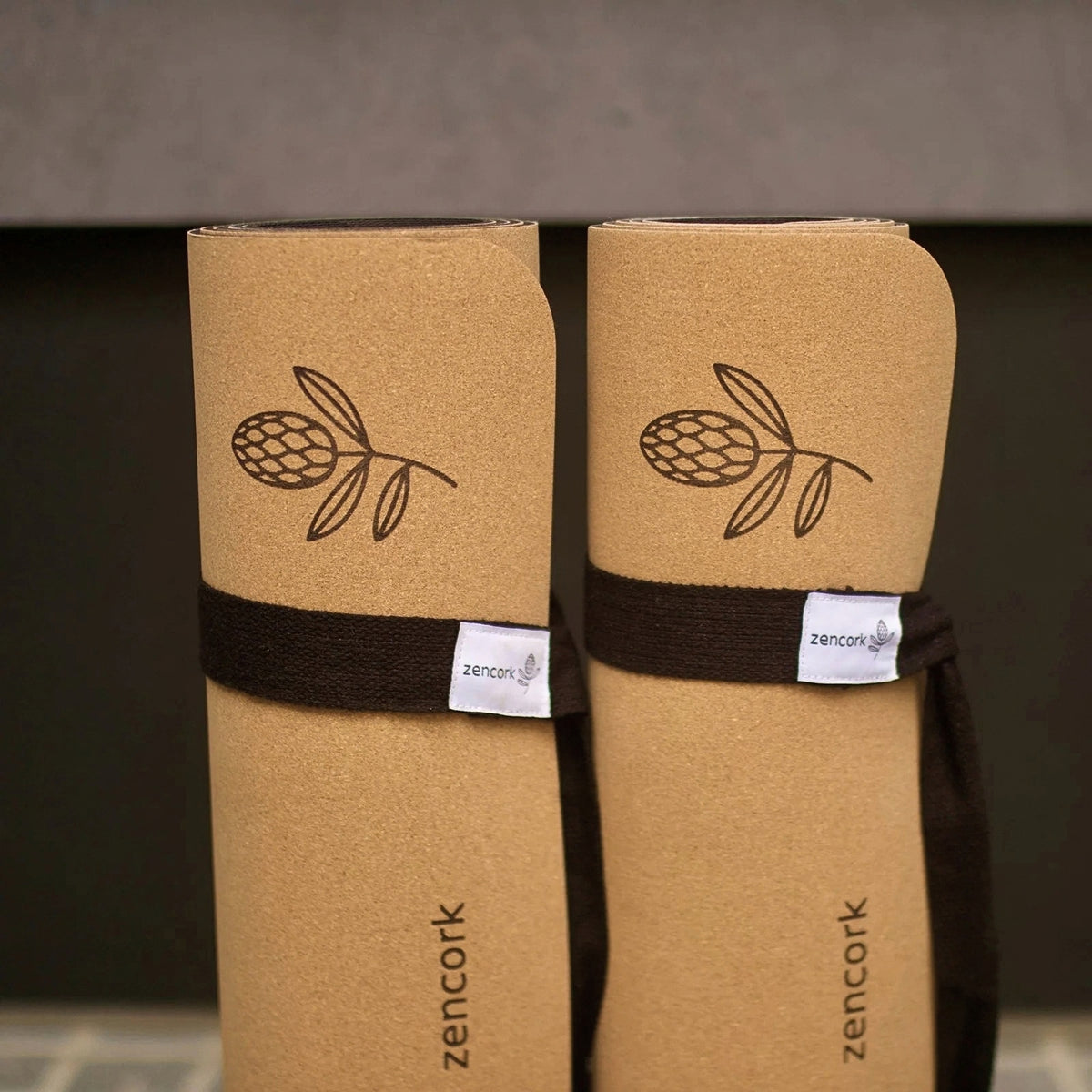 Two rolled tan cork yoga mats tied with dark brown bands, shown upright against a dark gray background. Each mat has a printed brown leaf logo and a small white tag with the brand name zencork.