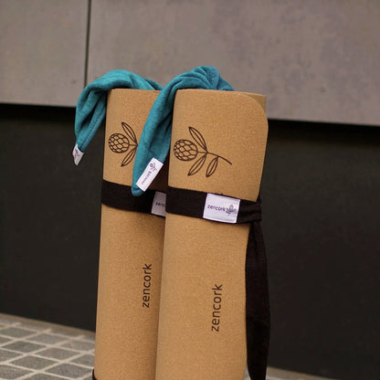 Two rolled tan yoga mats with black straps and a teal towel draped over them, standing against a dark gray wall. The mats are branded with the word 'zencork' and a simple leaf logo.