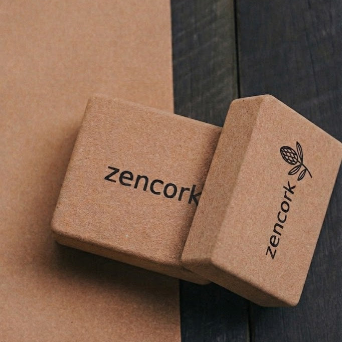 Two cork yoga blocks with the 'zencork' brand name and logo printed on them, resting on a dark wooden surface next to a brown textured background.