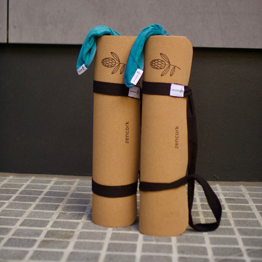 Two tan yoga mats with black straps and teal fabric loops, standing upright on a tiled surface against a dark wall.