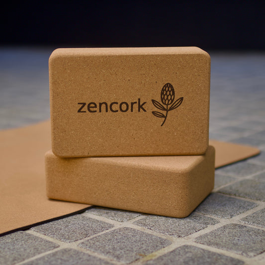Two natural cork yoga blocks stacked on a gray tiled surface with a brown cork mat underneath. The top block is printed with the brand name 'zencork' and a small botanical logo.