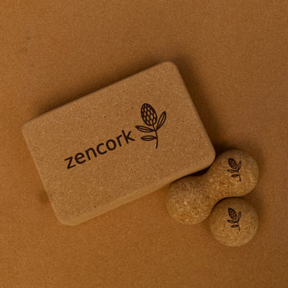 A cork yoga block with the brand name 'zencork' and a leaf logo, shown on a brown cork background with two small cork props.