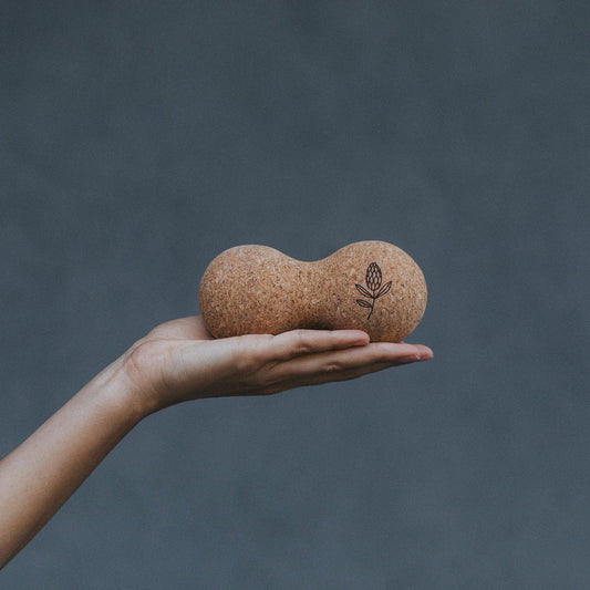 A hand holding a cork yoga prop shaped like a peanut with a small floral engraving, set against a dark gray background.