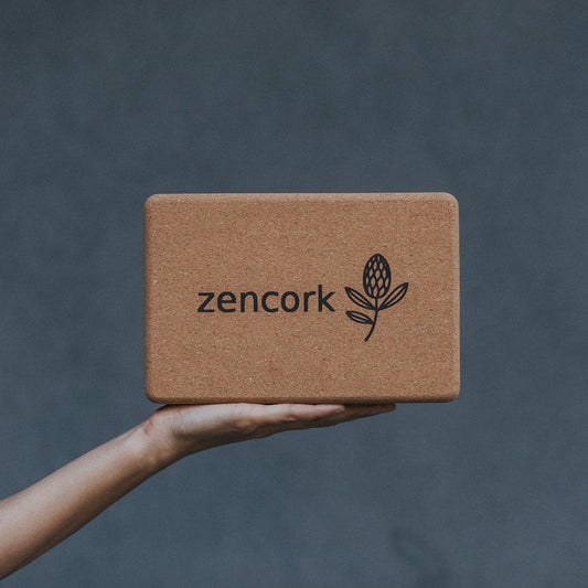 A cork yoga block with the brand name 'zencork' and a leaf logo printed on the front, held by a person's hand against a dark gray textured background.