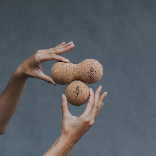 Two hands holding two cork massage rollers against a grey background