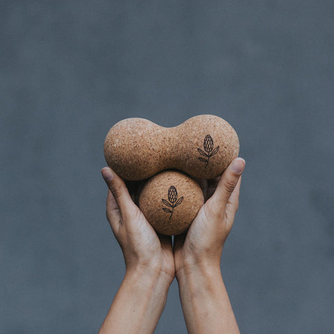 Two hands holding two cork yoga balls, with two engraved with a simple botanical leaf design, set against a dark textured gray background.