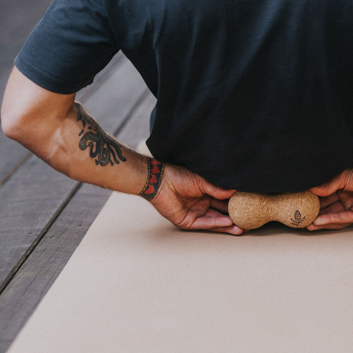 A close-up of a person using a small cork massage tool on their lower back, lying on a beige yoga mat on a wooden deck.