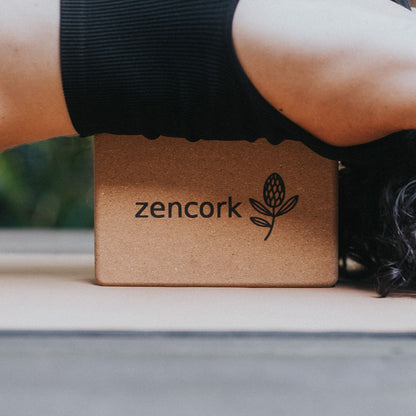 A cork yoga block with the brand name 'zencork' and a leaf logo printed on it, positioned under a person's arm wearing a black ribbed sleeve.