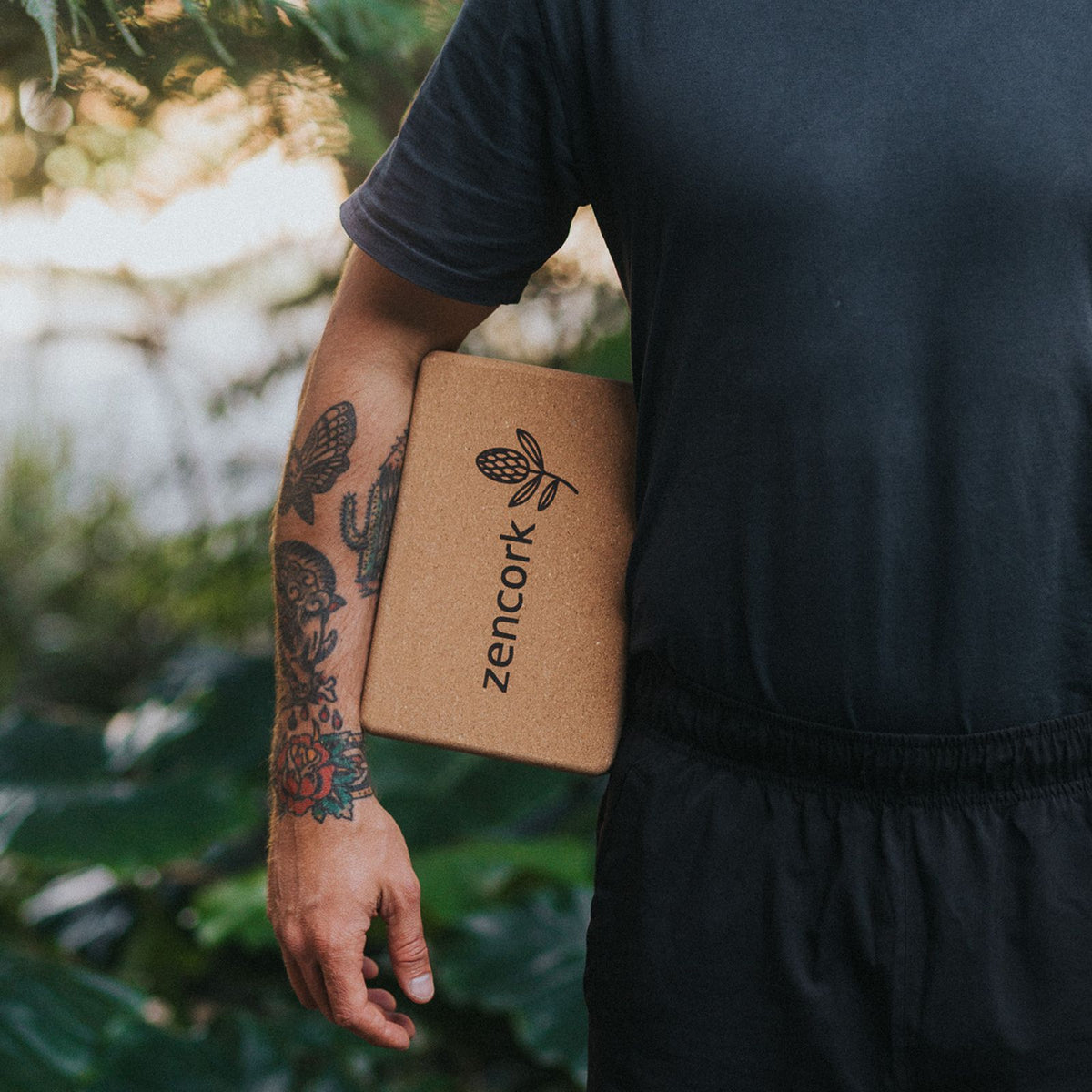 A person outdoors holding a tan cork yoga block branded with 'zencork' in black text. The person is wearing a dark gray t-shirt and black shorts, with a blurred background of green foliage.