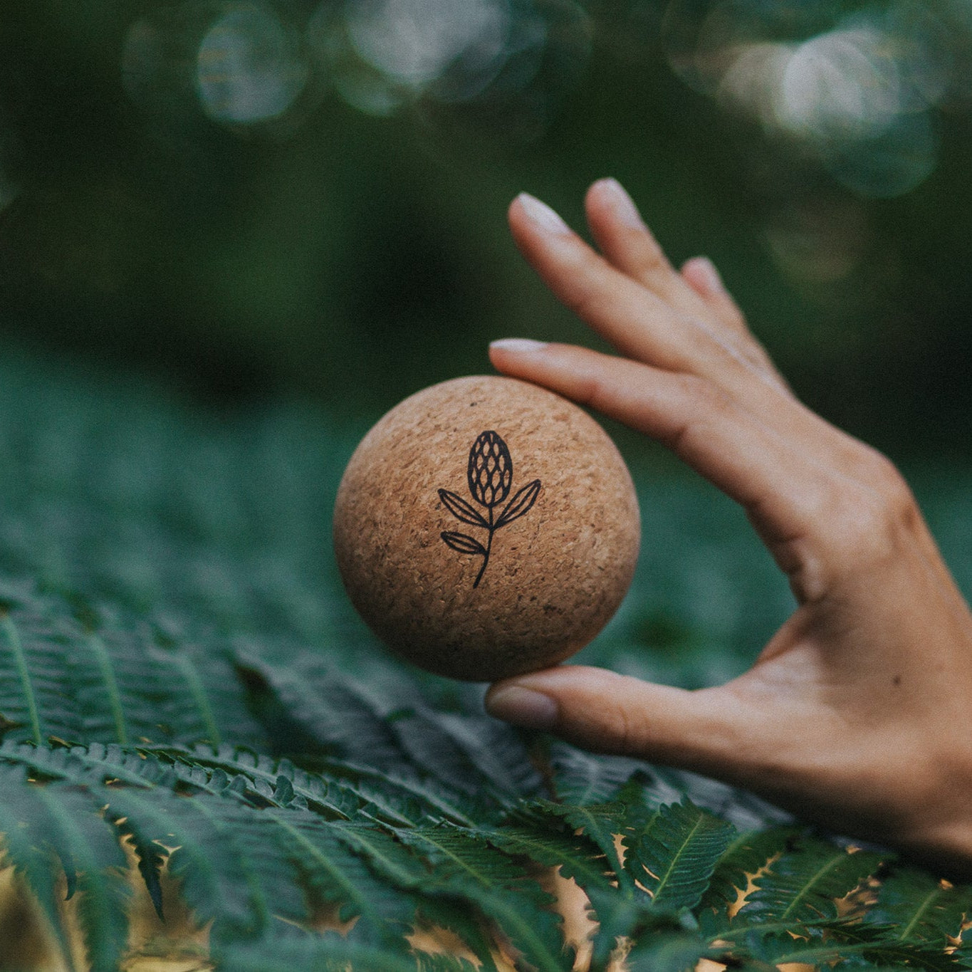 Hand holding a cork ball with a leaf design against a blurred green foliage background