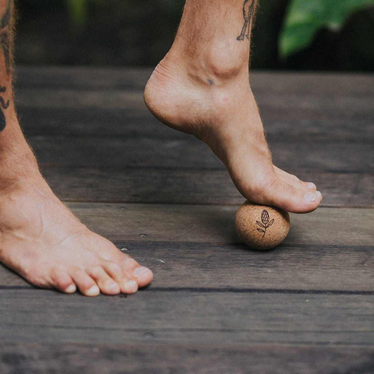 A close-up of a person's bare feet using a small cork massage ball on a dark wooden deck. The cork ball has a laser engraved banksia flower.