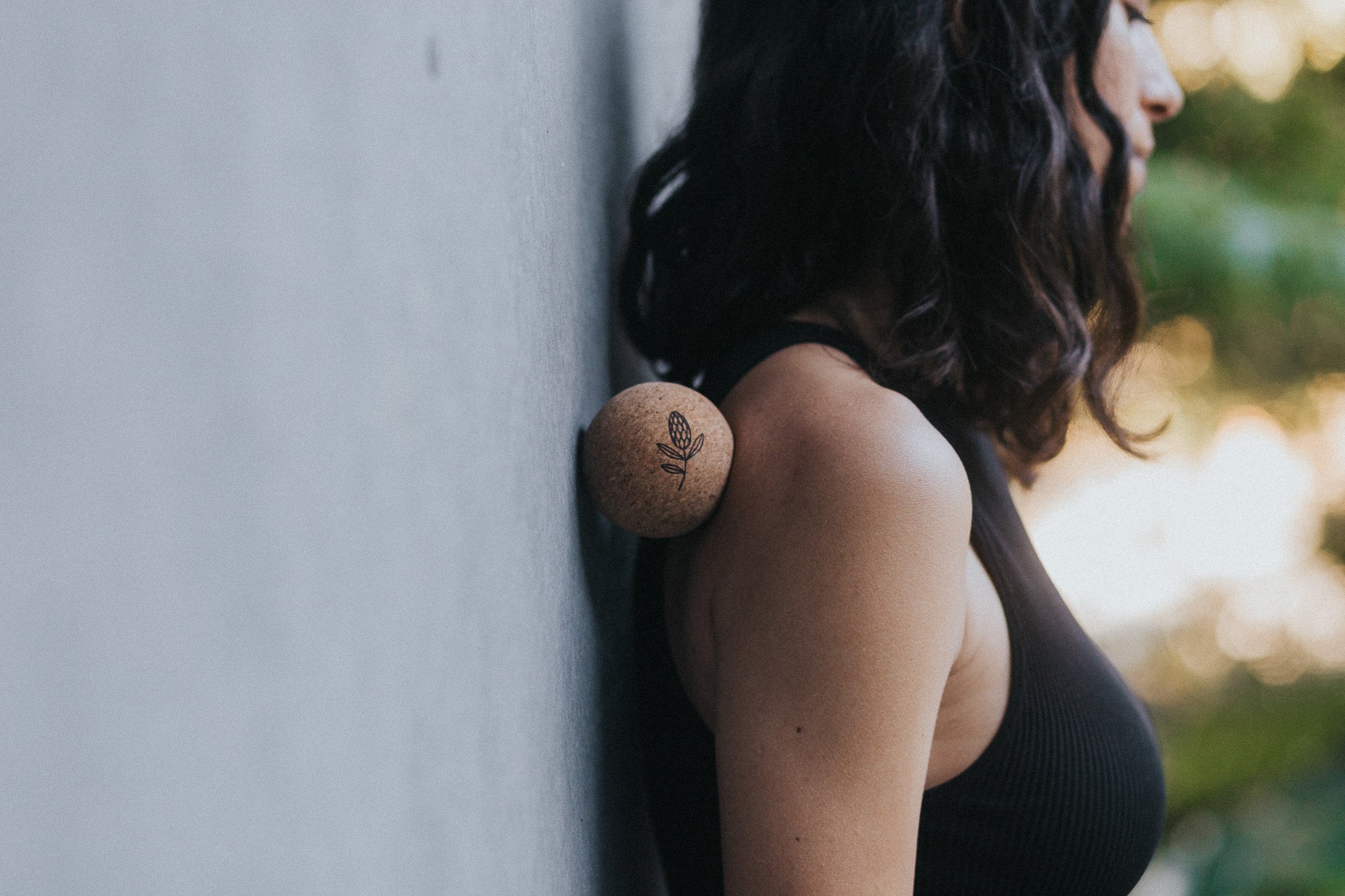 Person wearing a black sleeveless top with a cork yoga ball against her shoulder, leaning against a wall.