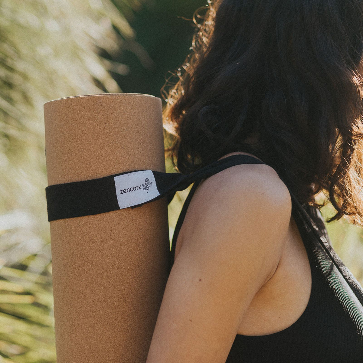 A person outdoors carrying a tan yoga mat over their shoulder with a black strap across their upper back. The strap has a white label that reads 'ZenCork'.