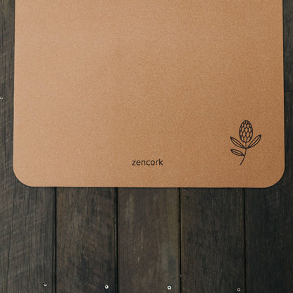A tan cork yoga mat with rounded corners, featuring the brand name 'zencork' and a small botanical line drawing in the bottom right corner, displayed on a dark wooden plank surface.