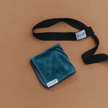 A folded teal blue-green cloth with a small white tag, placed beside a black strap with a matching white tag, on a warm tan background.