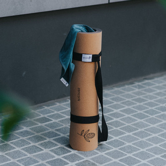 A rolled tan yoga mat with black straps and a teal towel draped over the top, standing on a gray tiled floor against a dark gray wall. The mat has a small Banksia logo and the brand name 'ZenCork' printed vertically.