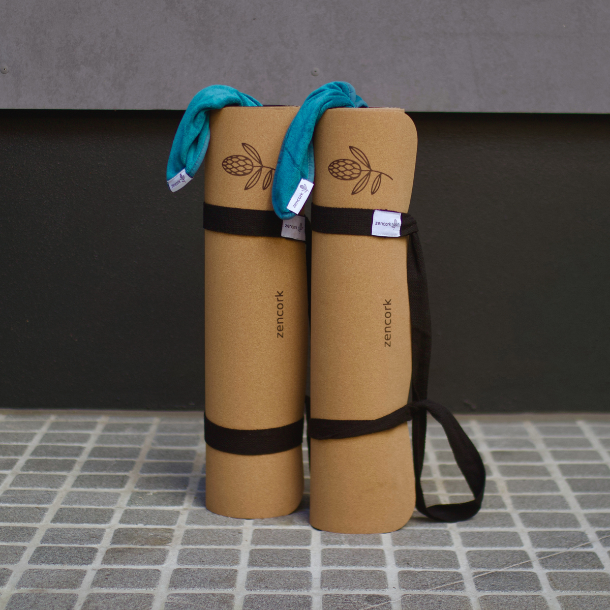 Two rolled tan yoga mats with black straps and a teal towel draped over the top, standing on a tiled surface against a dark wall. The mats are branded ZENCOOK.