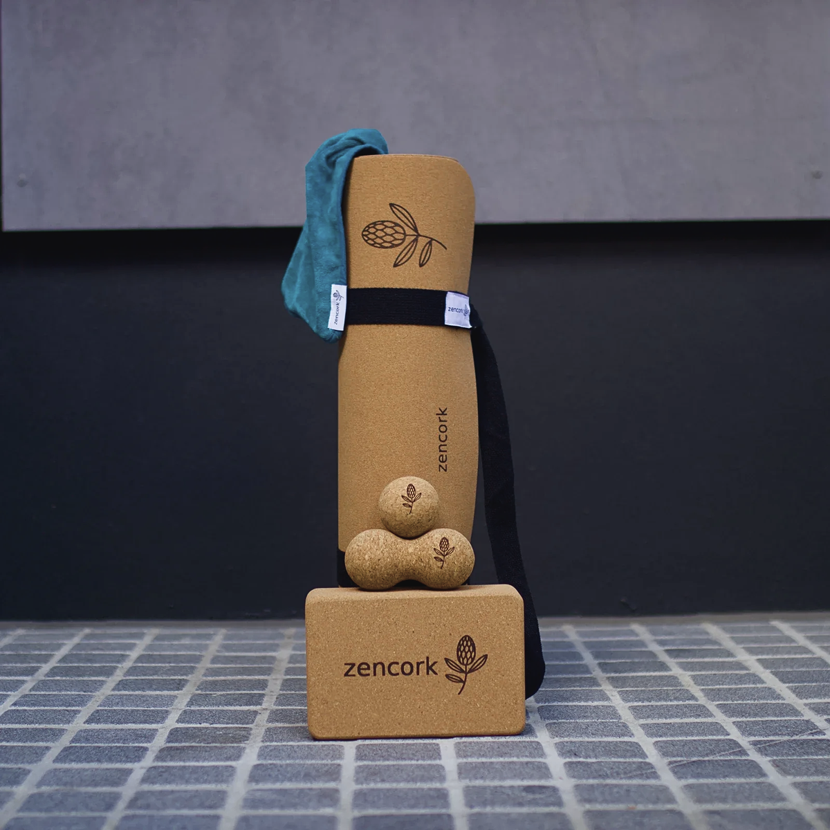 A cork yoga set including a rolled tan cork mat with a teal fabric cover, three small cork massage balls, and a tan cork block, all branded 'zencork' with a botanical logo, arranged on a tiled floor against a dark wall.